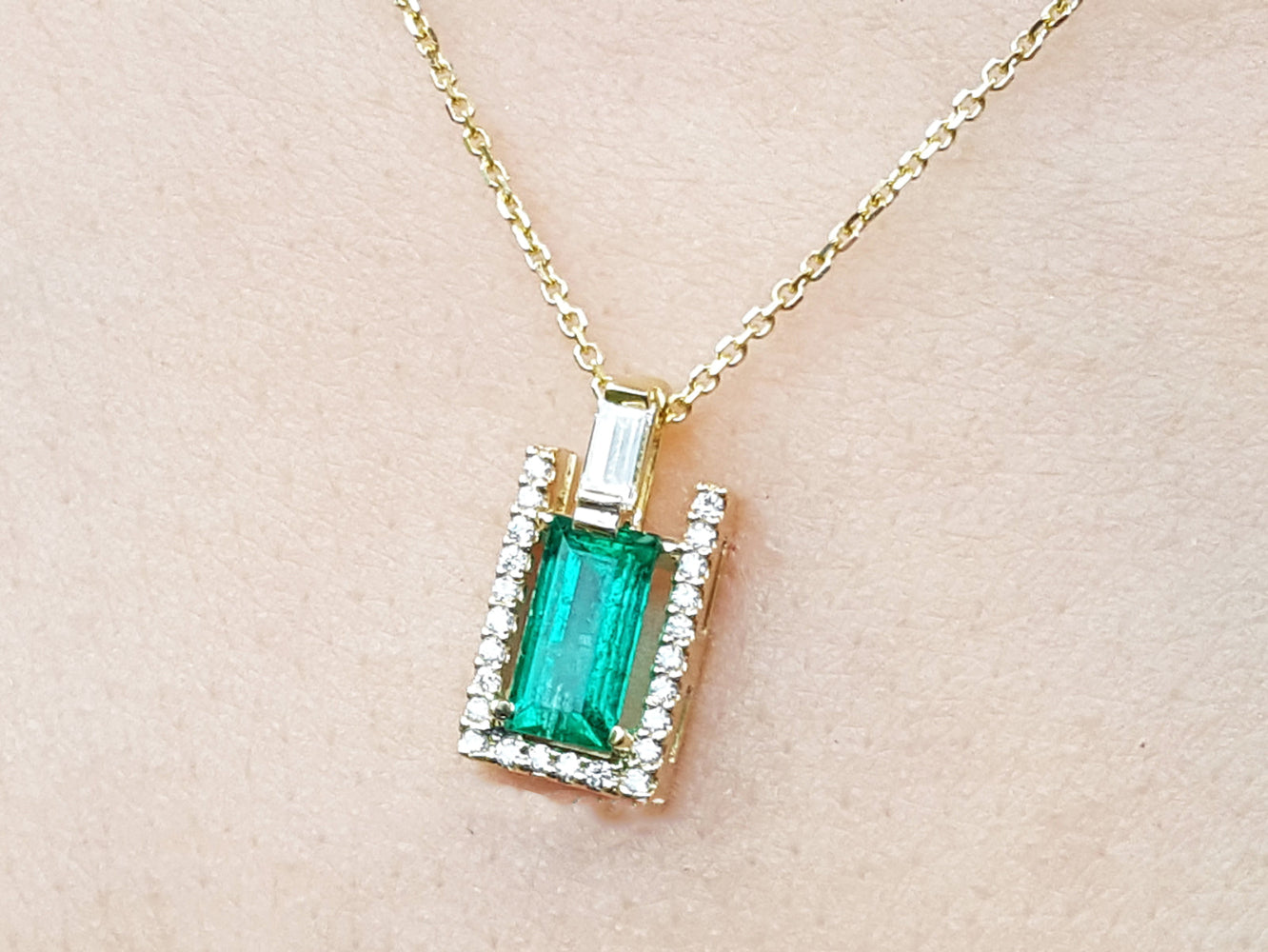 Gold necklace with a baguette cut emerald pendant on a beige skin background.