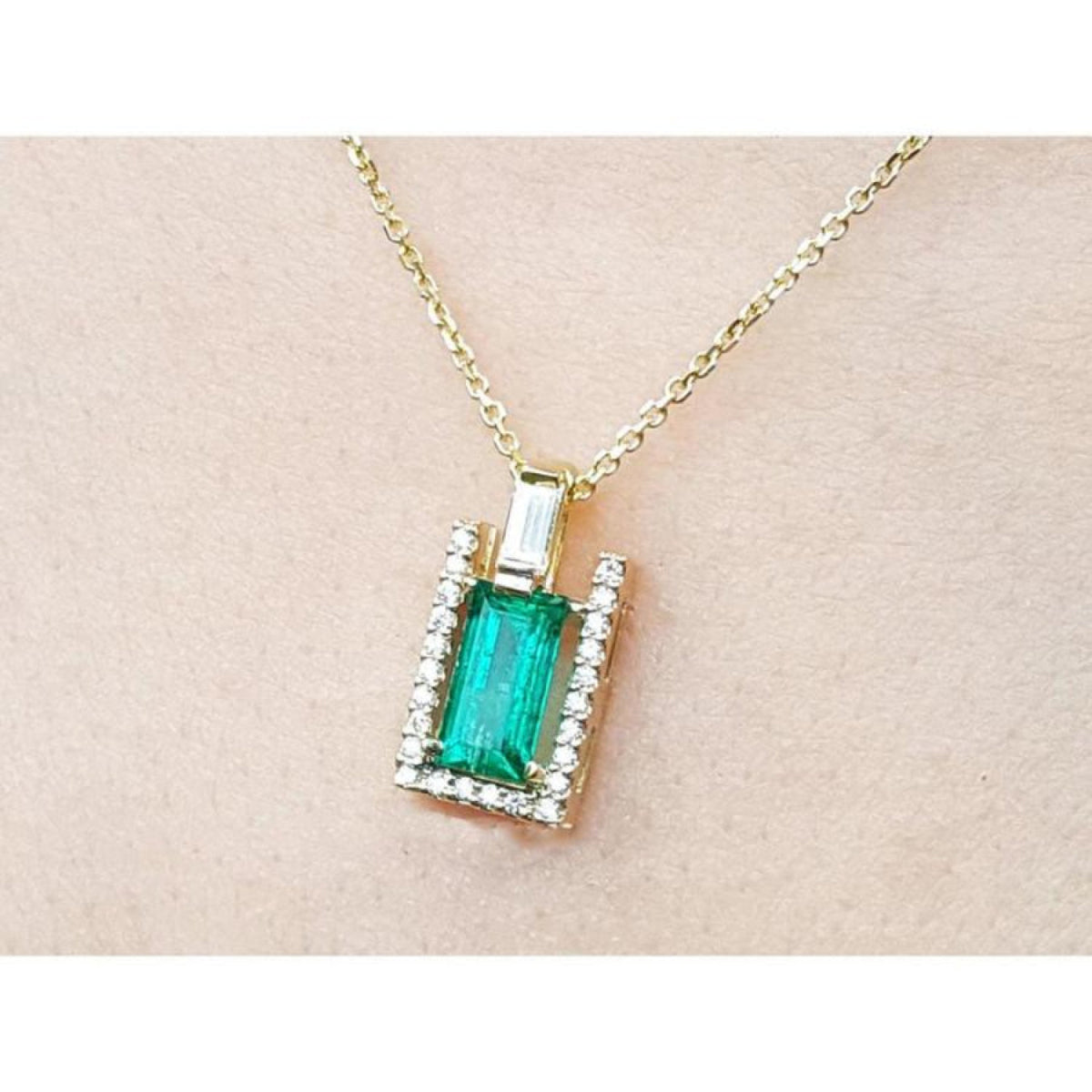 Gold necklace with a baguette cut emerald pendant on a beige skin background.
