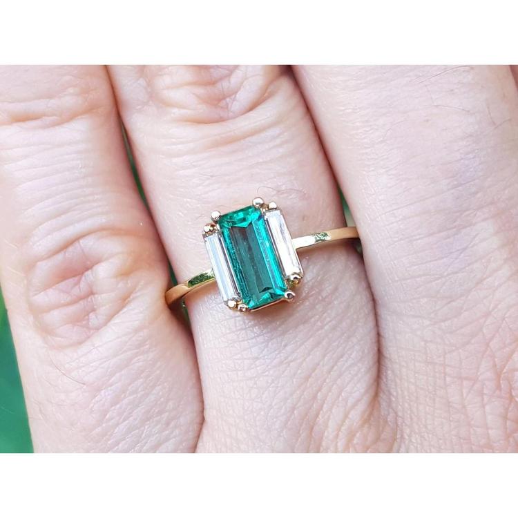 A baguette cut emerald ring with side diamonds crafted in 18k yellow gold on a finger.