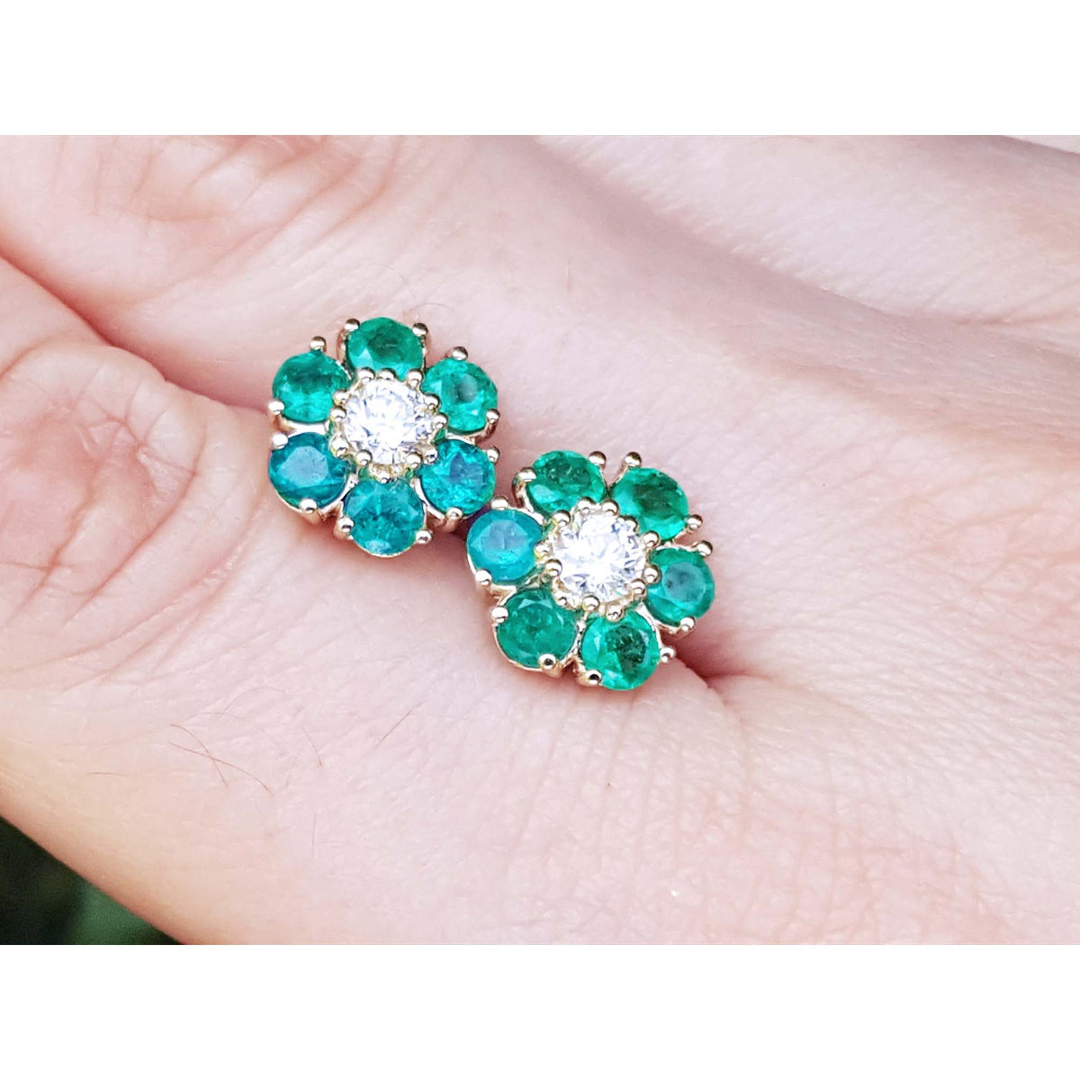 Pair of emerald and diamond cluster stud earrings held between fingers.