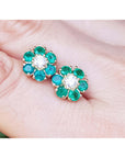 Pair of emerald and diamond cluster stud earrings held between fingers.