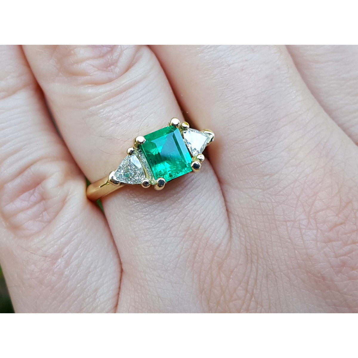A square natural emerald with side trillion diamonds in 18k yellow gold on a hand.