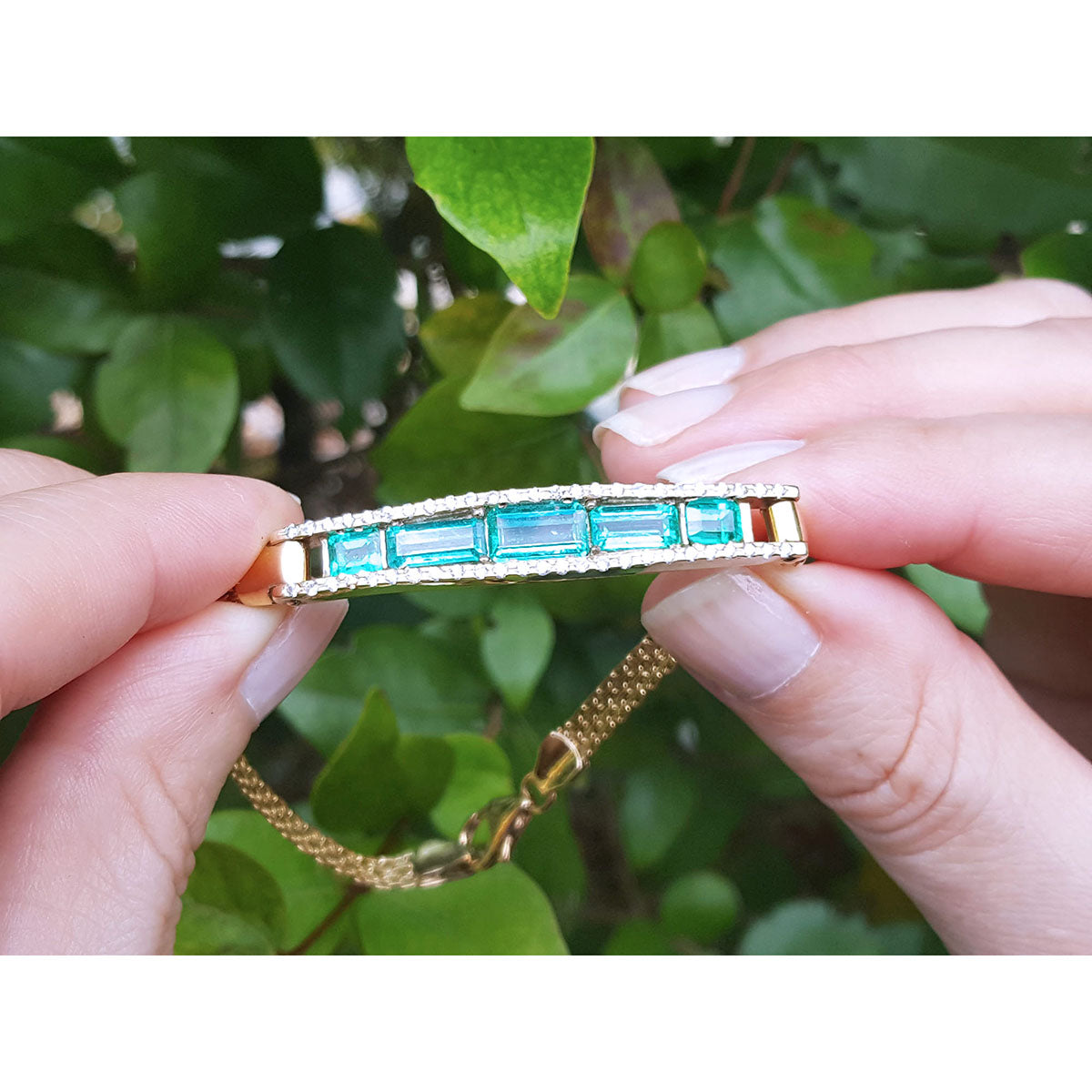 A yellow gold bracelet with emeralds and side diamonds held by hands against a leafy background.