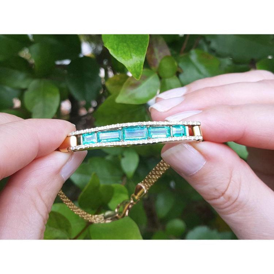 14K gold bracelet with five vibrant bluish-green emeralds and surrounding diamonds, held against green leaves.