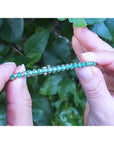 Hands holding between fingers a colombian emerald tennis bracelet against a green leafy background.