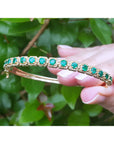 Gold bracelet with round cut emeralds held by a hand between fingers against a green leafy background.