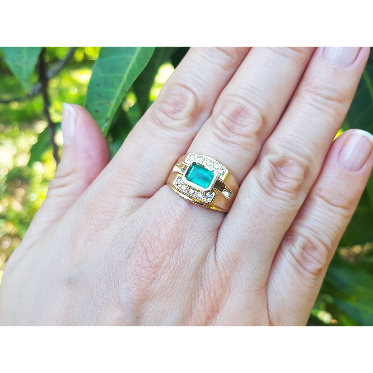 A hand wearing a yellow gold mens emerald ring against a green leafy background.