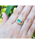 A hand wearing a yellow gold mens emerald ring against a green leafy background.
