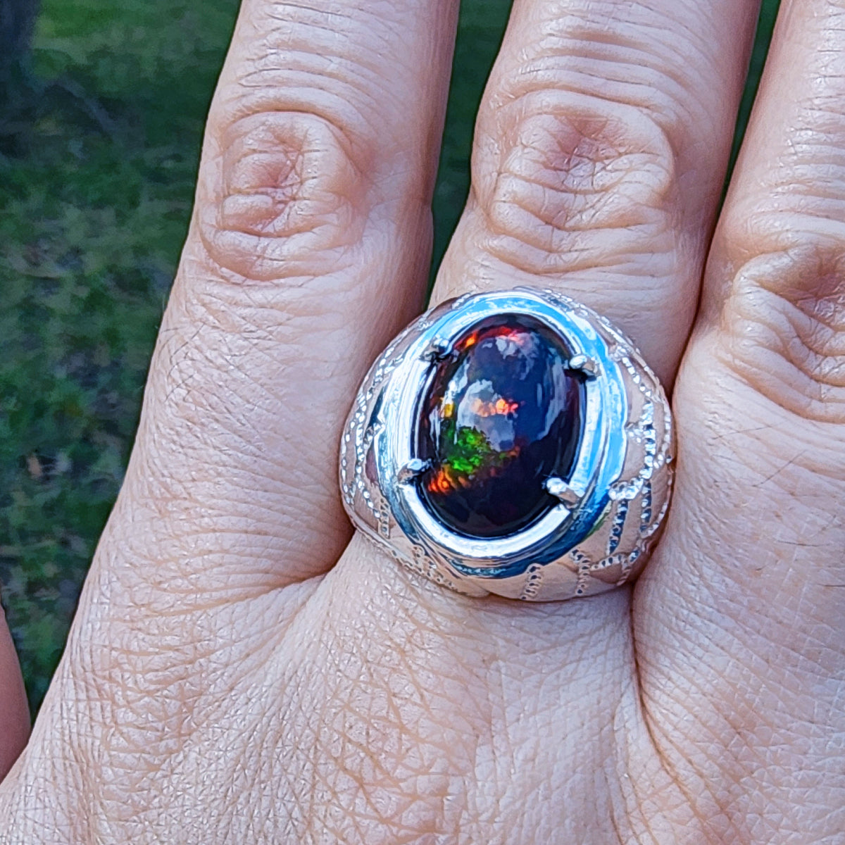 Men's silver ring with a colorful black opal held between fingers against a blurred green background