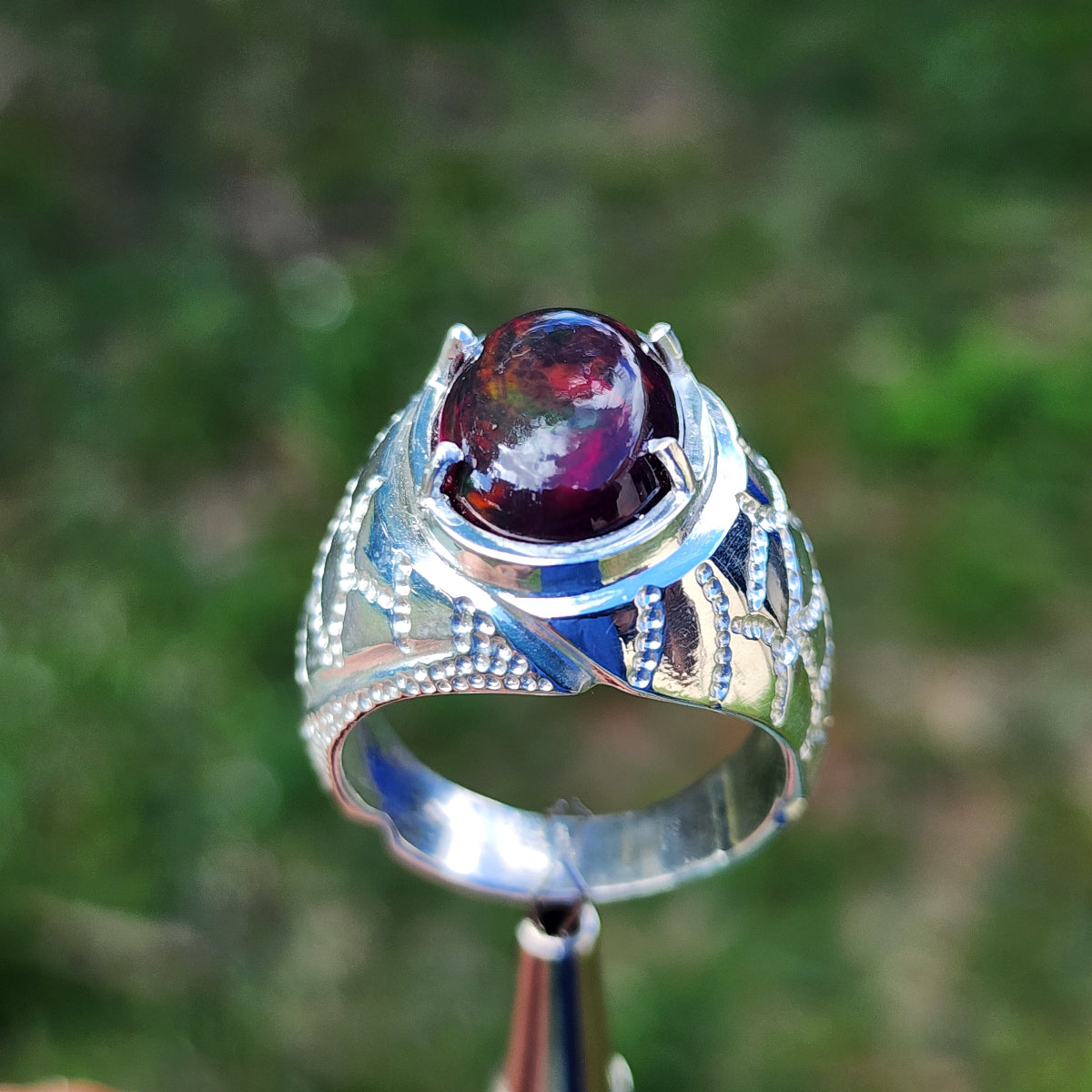 Men's silver ring with a purple black opal on a stand against a blurred green background