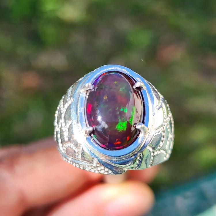 Silver men's ring with a colorful black opal held by a hand against a blurred green background.