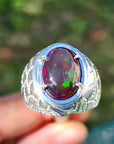 Silver men's ring with a colorful black opal held by a hand against a blurred green background.