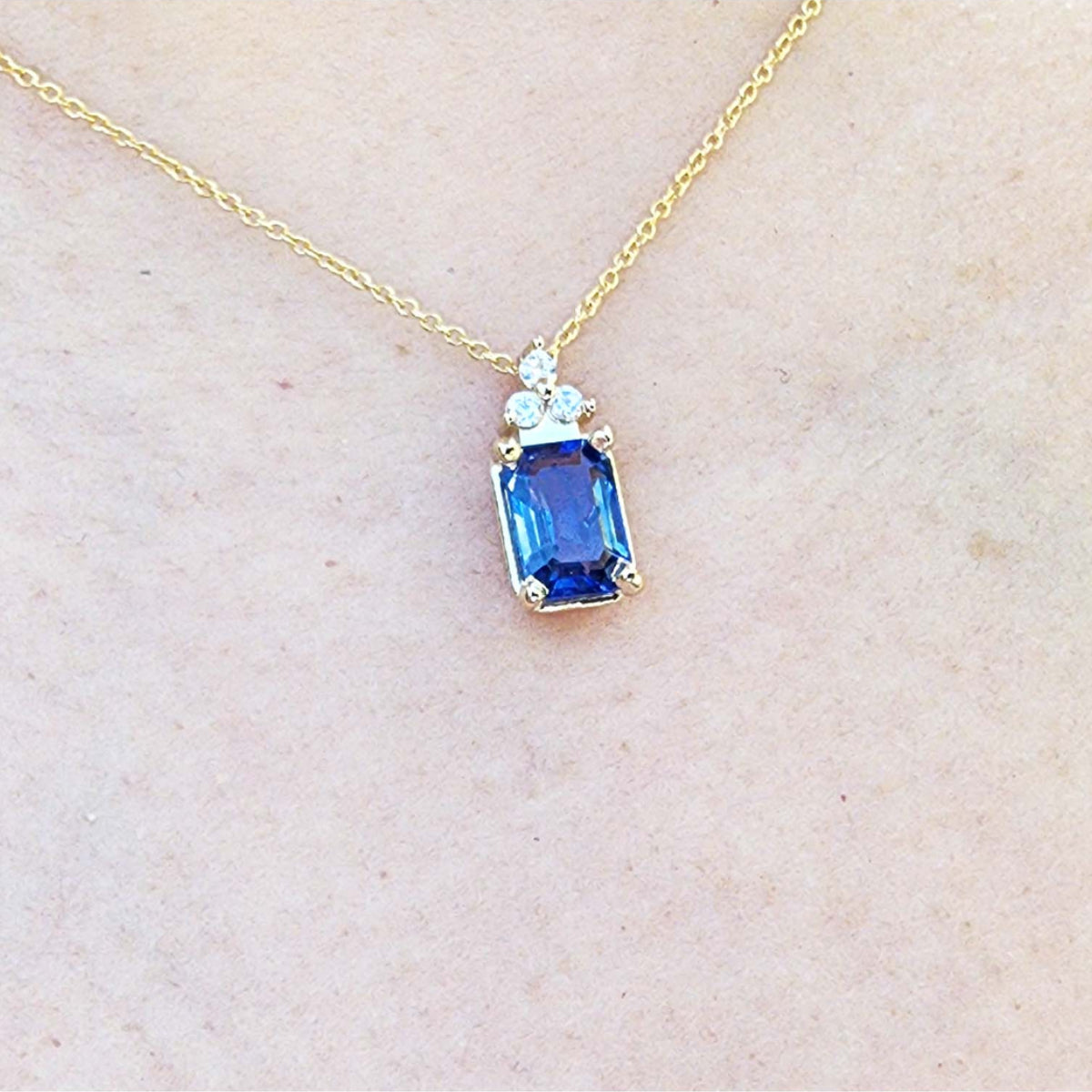 Gold necklace with a blue genuine sapphire pendant on a light beige skin background.