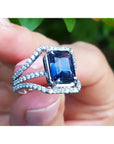 18k white gold ring with a natural blue sapphire and diamond accents held between finger against a blurred green background