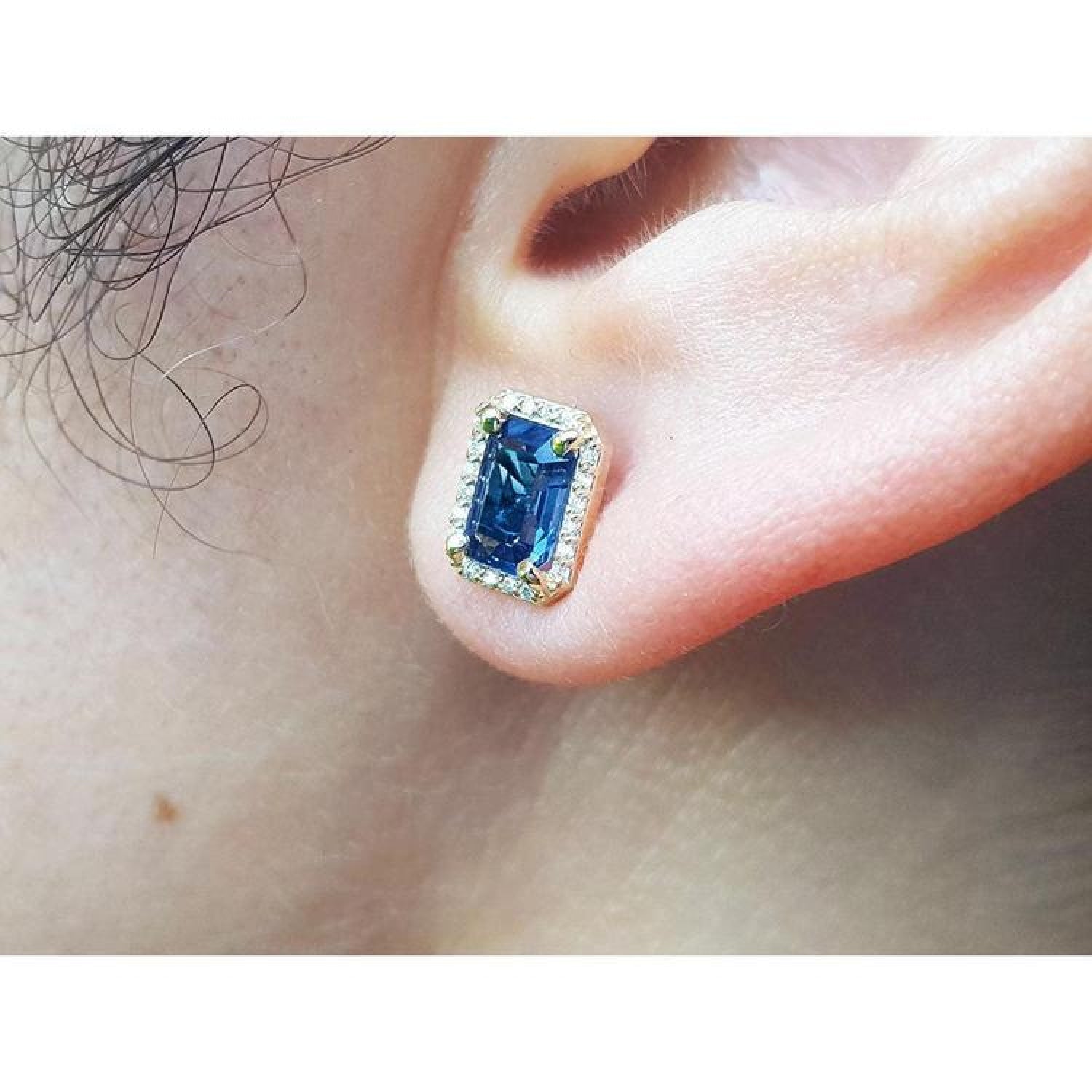 Emerald cut blue sapphire stud earring with halo diamonds in 14K yellow gold on an ear.