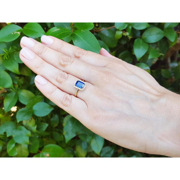 Hand wearing a yellow gold ring with a blue natural sapphire against a green leafy background