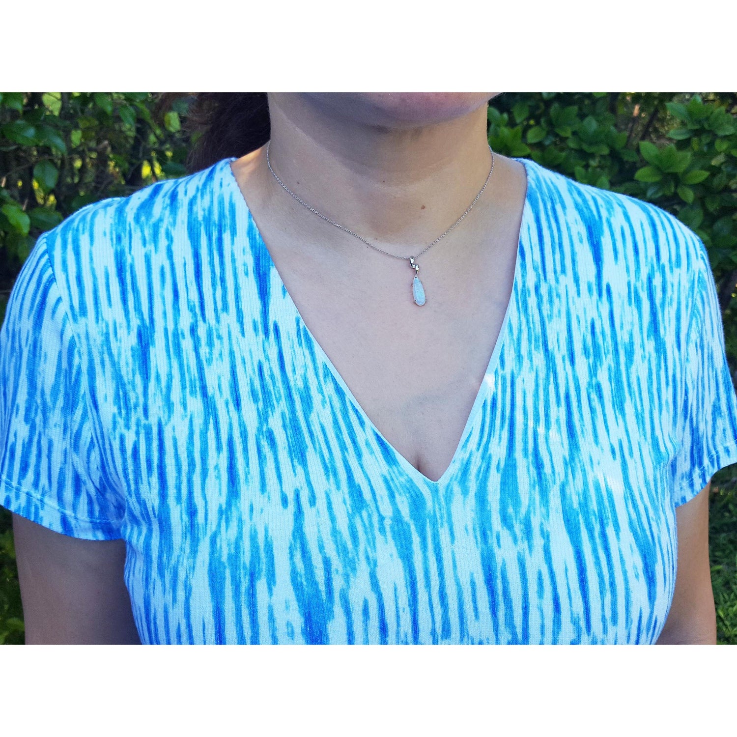Person wearing an opal pendant in a blue and white tie-dye shirt with a blurred green background.