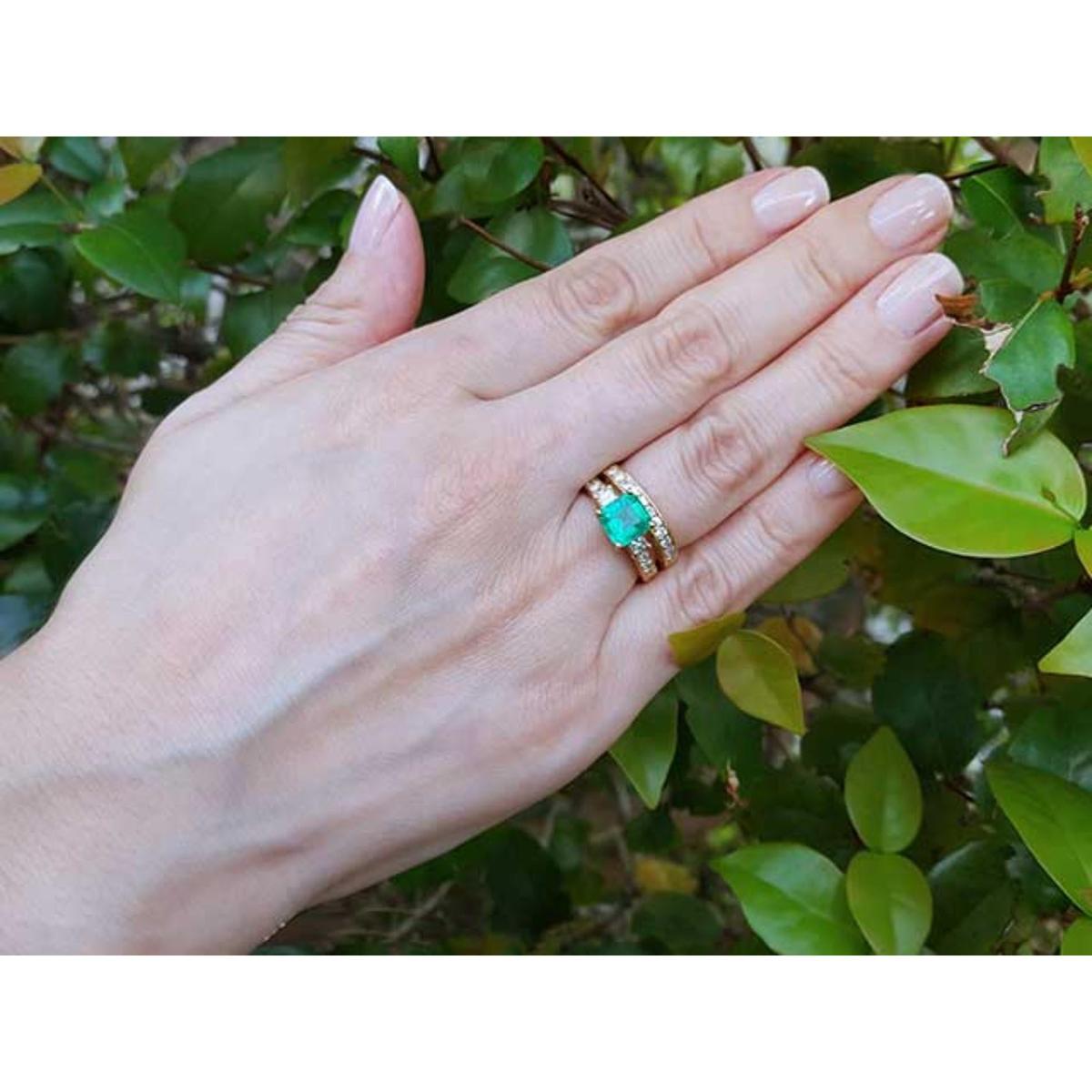 Hand wearing a ring with an emerald stone with matching wedding band against a green leafy background.