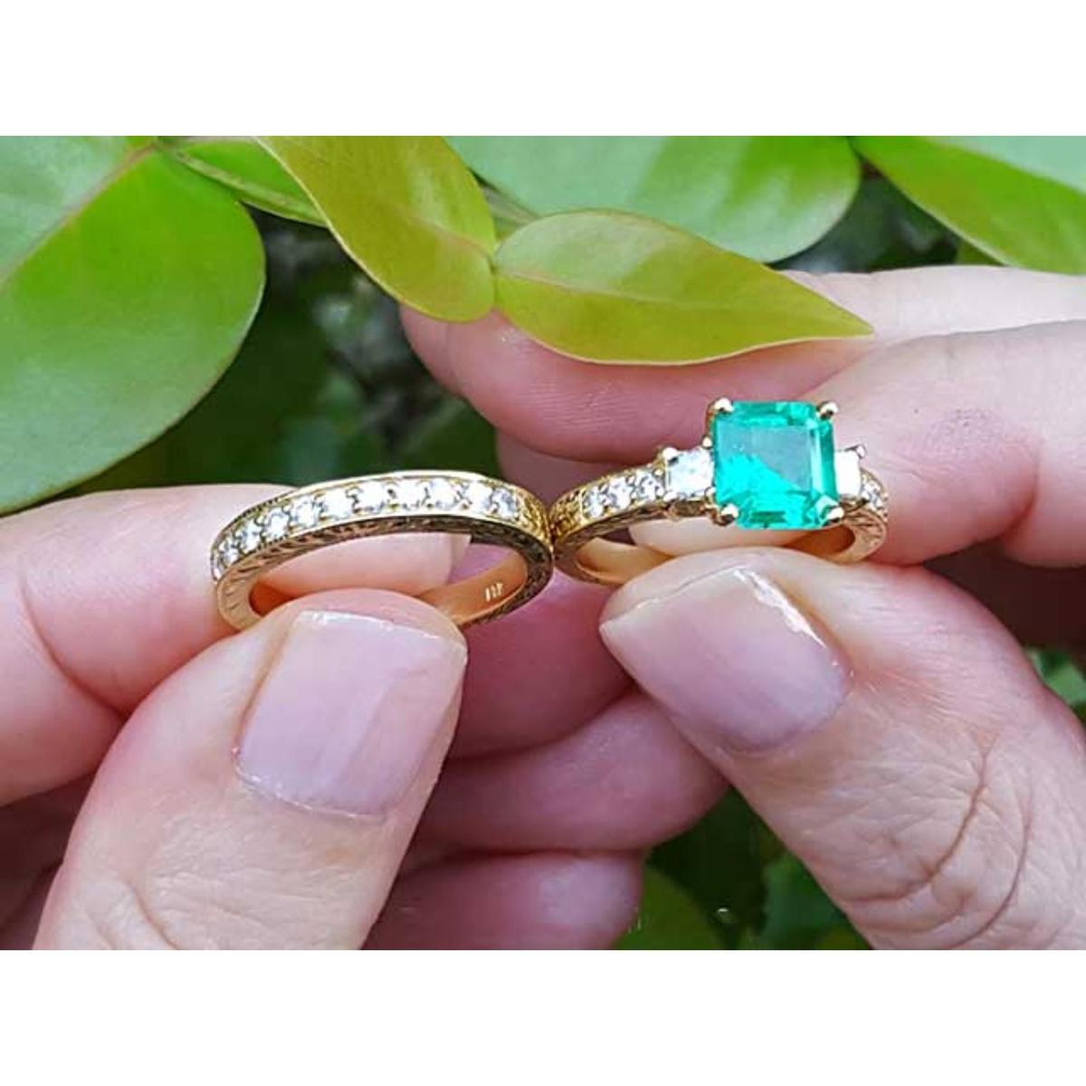 Both, emerald ring and matching wedding band held between fingers against a green leafy background.