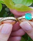 Both, emerald ring and matching wedding band held between fingers against a green leafy background.