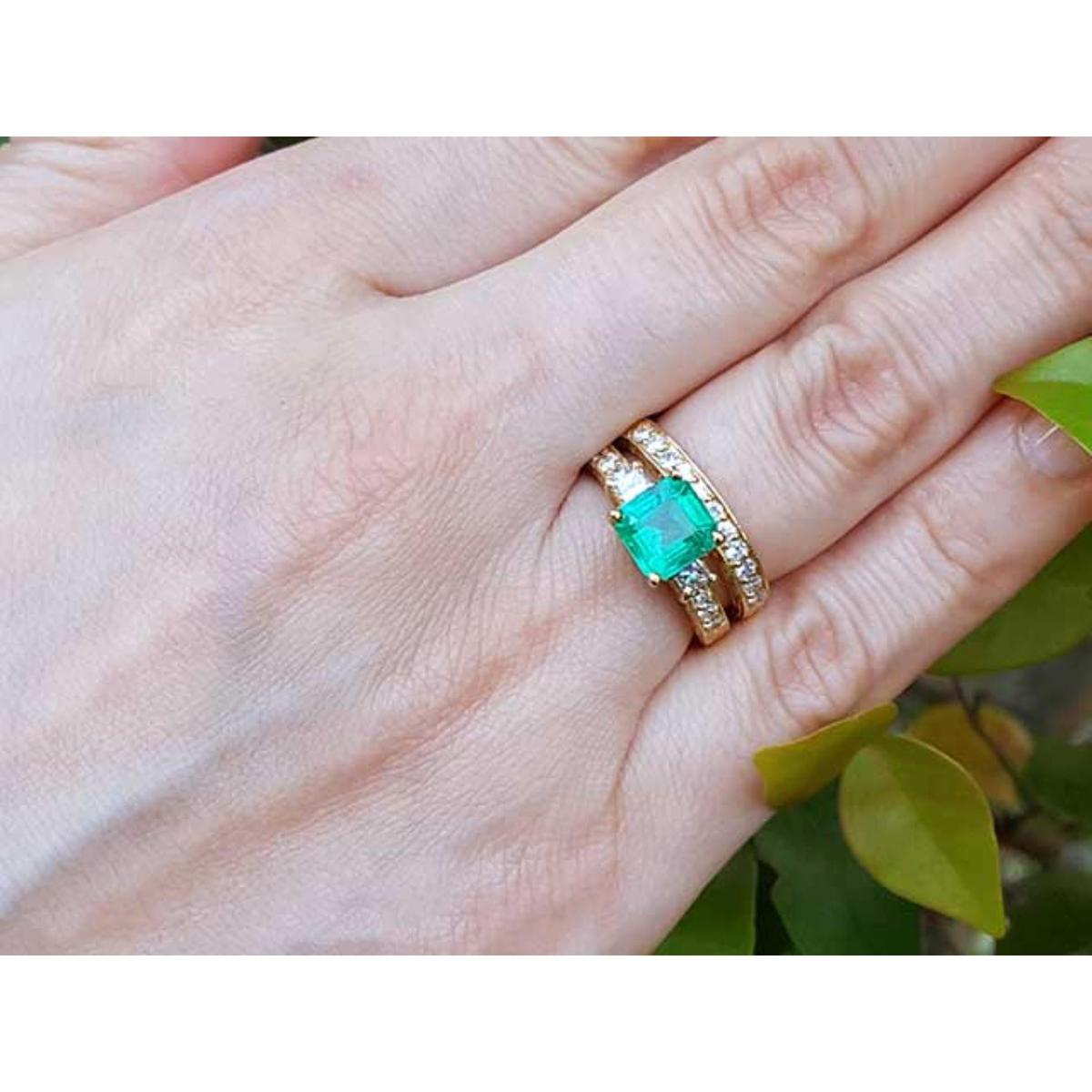 The top view of a yellow gold emerald ring with matching wedding band on a hand, with leaves in the background.
