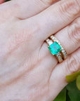 The top view of a yellow gold emerald ring with matching wedding band on a hand, with leaves in the background.