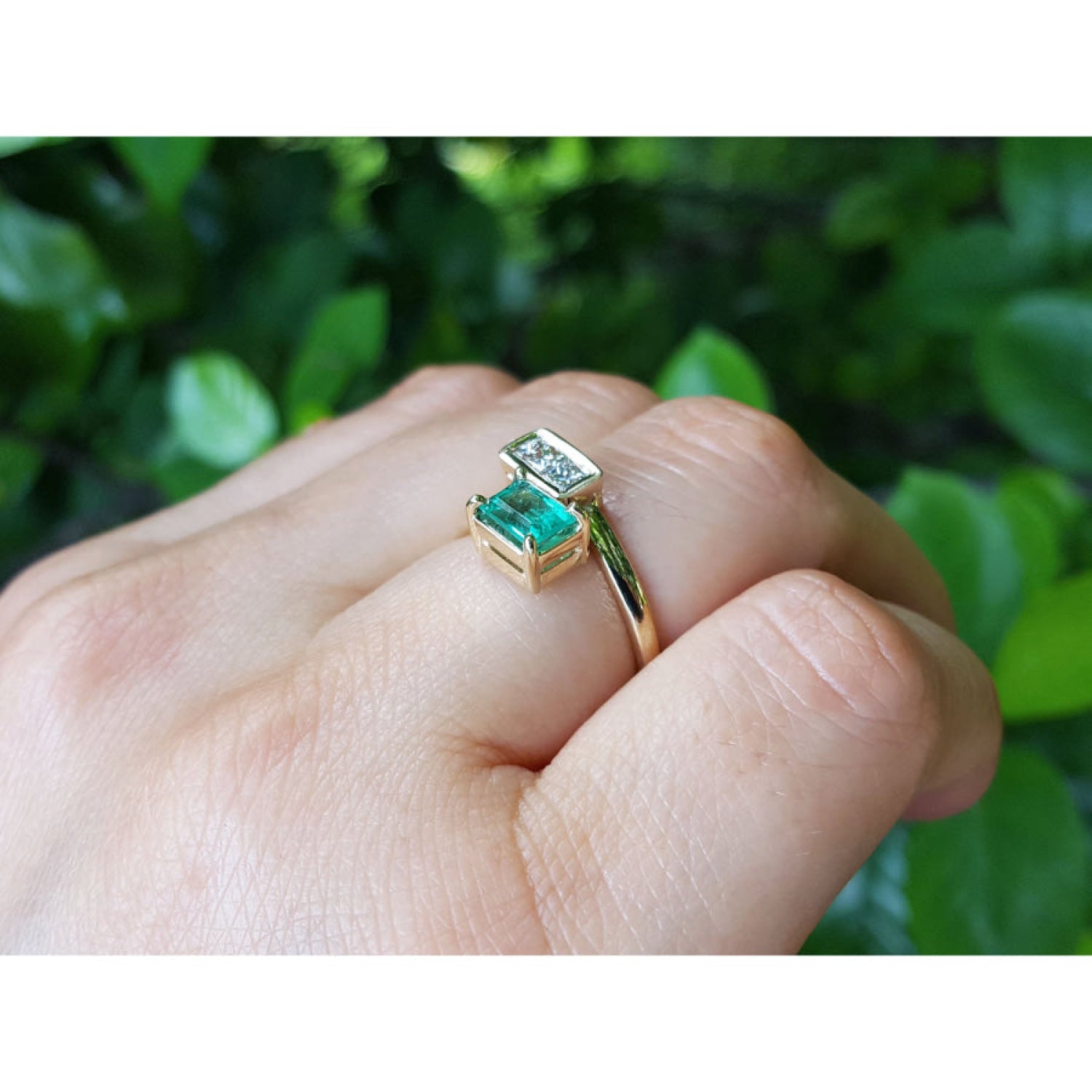 A 18k gold you and me or toi et moi emerald and diamond ring, worn on a person's finger against a background of green foliage.