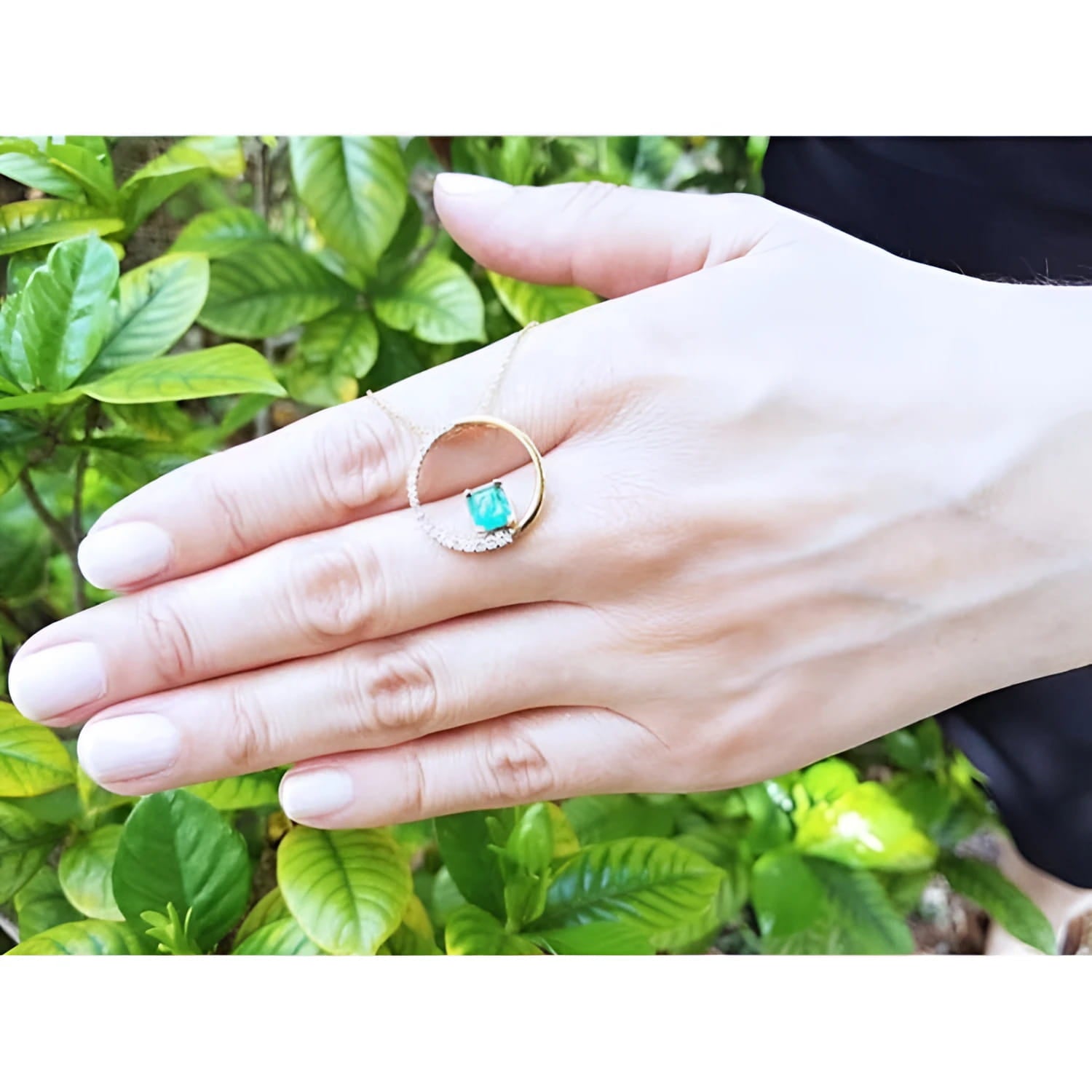 14K gold Circle of Life emerald pendant with natural diamonds, worn on a hand against green leaves.