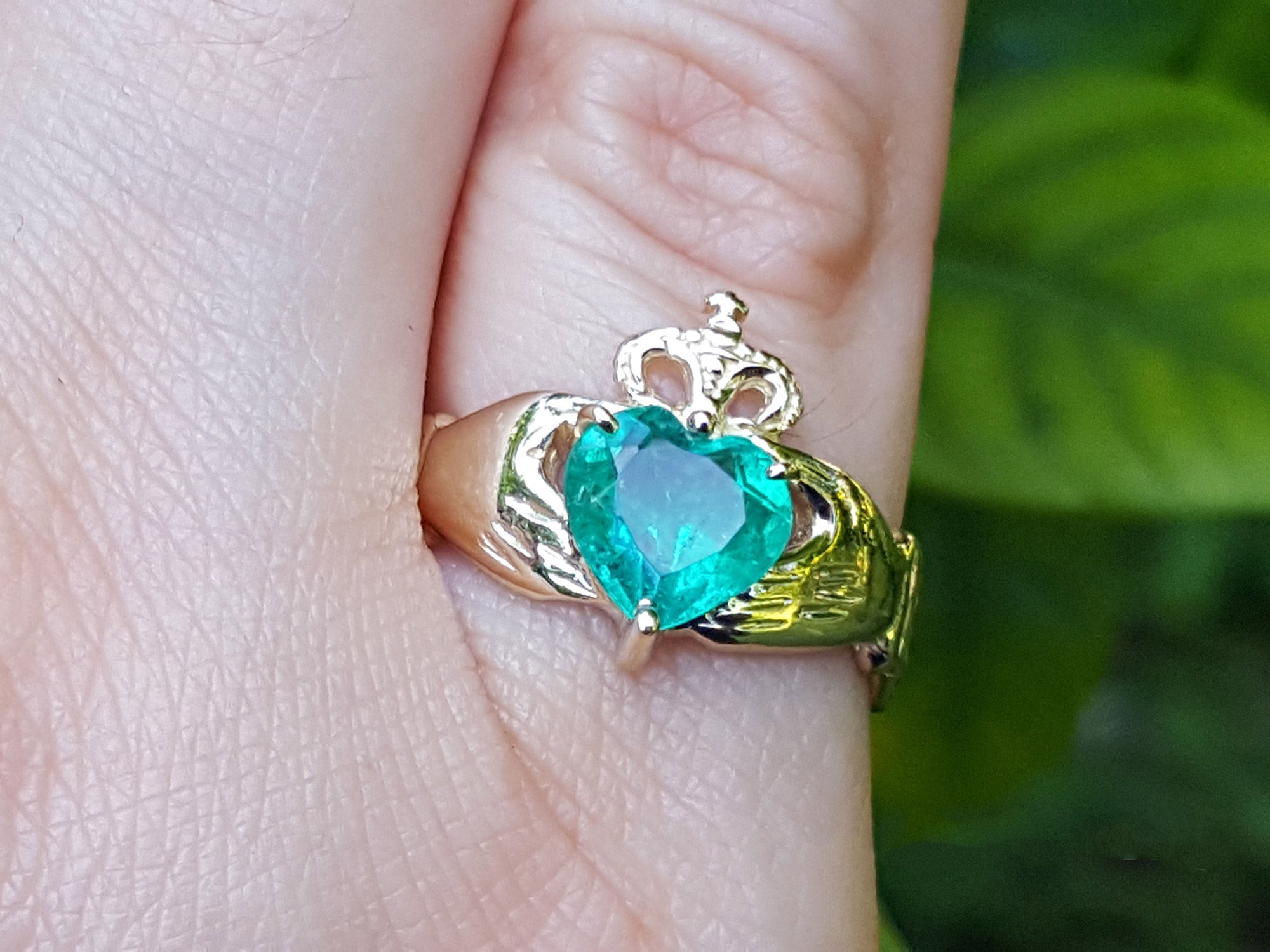 A yellow gold heart emerald claddagh ring on a finger against a blurry green background.