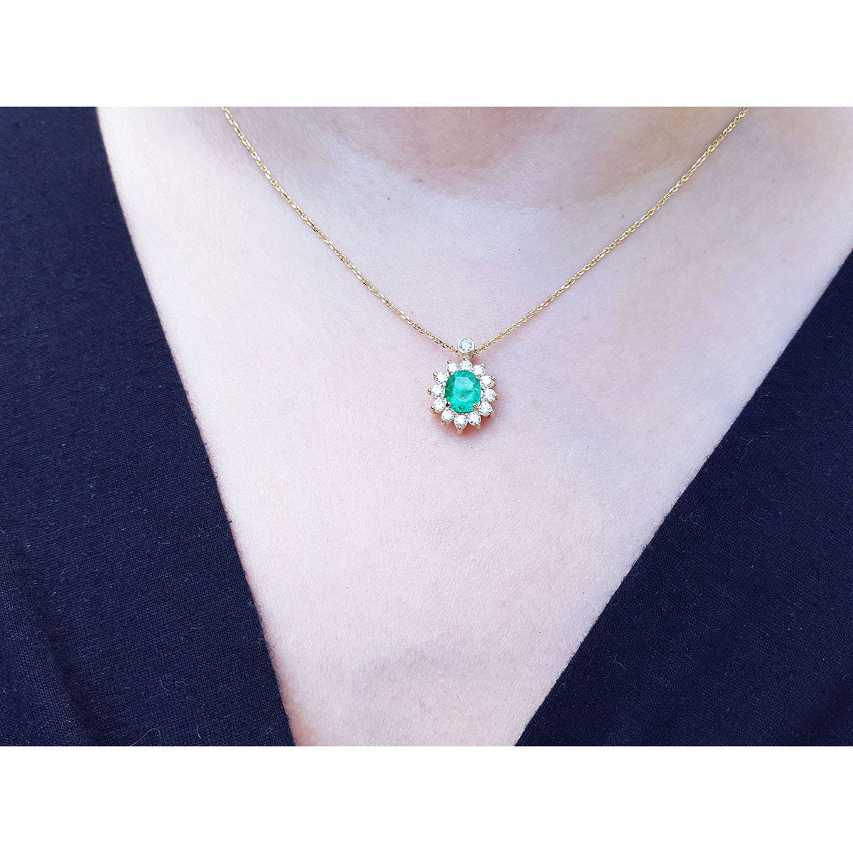 A close sight of a gold necklace with a oval Muzo emerald gemstone pendant on a person wearing a dark blue top.