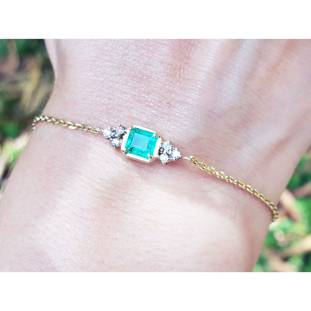 Gold bracelet with a square colombian emerald on a wrist against a blurred natural background.