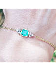 Gold bracelet with a square colombian emerald on a wrist against a blurred natural background.