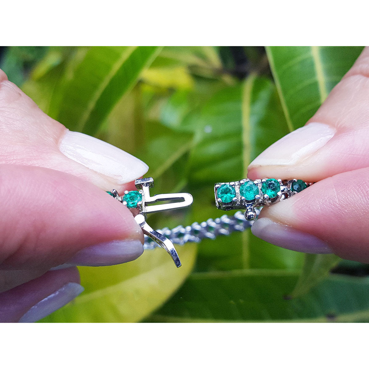 Close-up of hands holding a white gold tennis bracelet with round cut emeralds against a leafy background.
