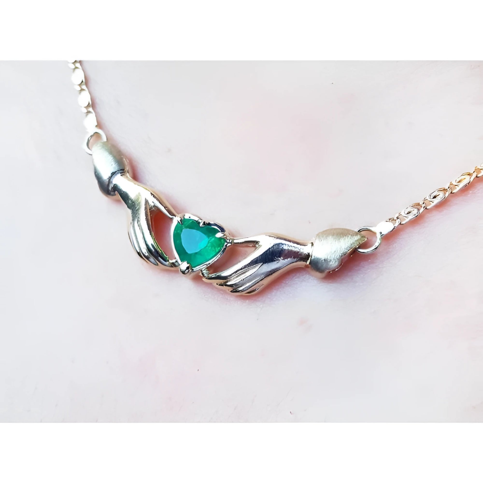 14K yellow gold Claddagh necklace with a heart-cut emerald held by two hands, worn on a neck.