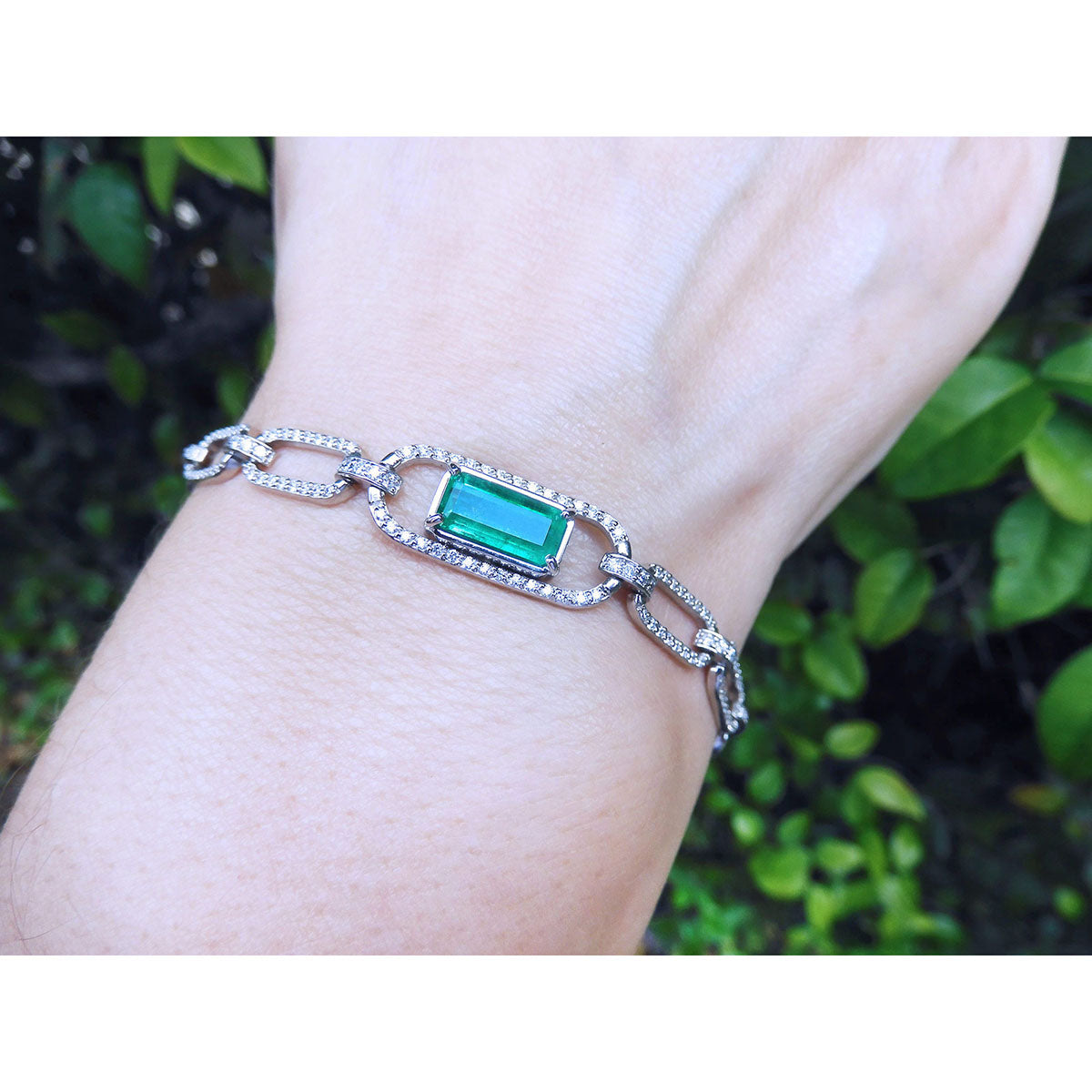 A white gold paperclip style bracelet with emerald on a wrist against a natural background.