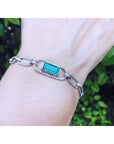 A white gold paperclip style bracelet with emerald on a wrist against a natural background.