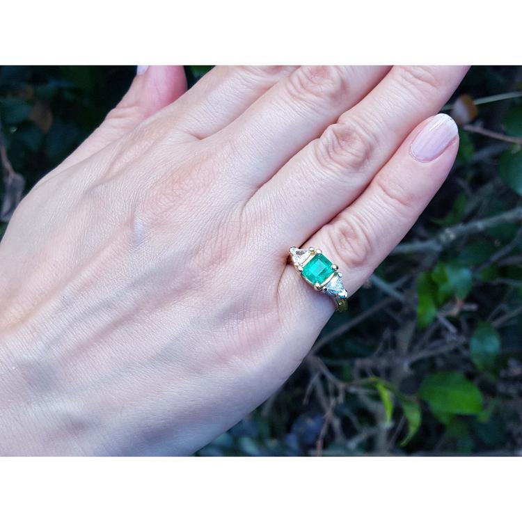 Hand wearing a 18k yellow gold ring with a square colombian emerald and trillion diamonds against a natural background.