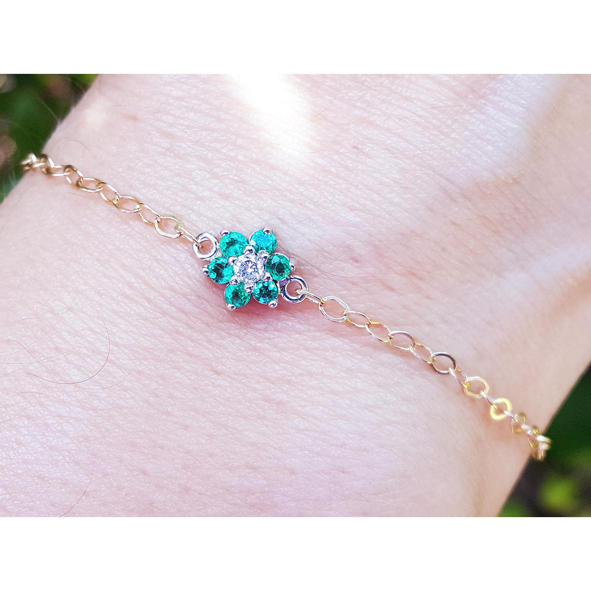 Gold bracelet with a emerald flower-shaped clasp on a wrist against a blurred natural background.