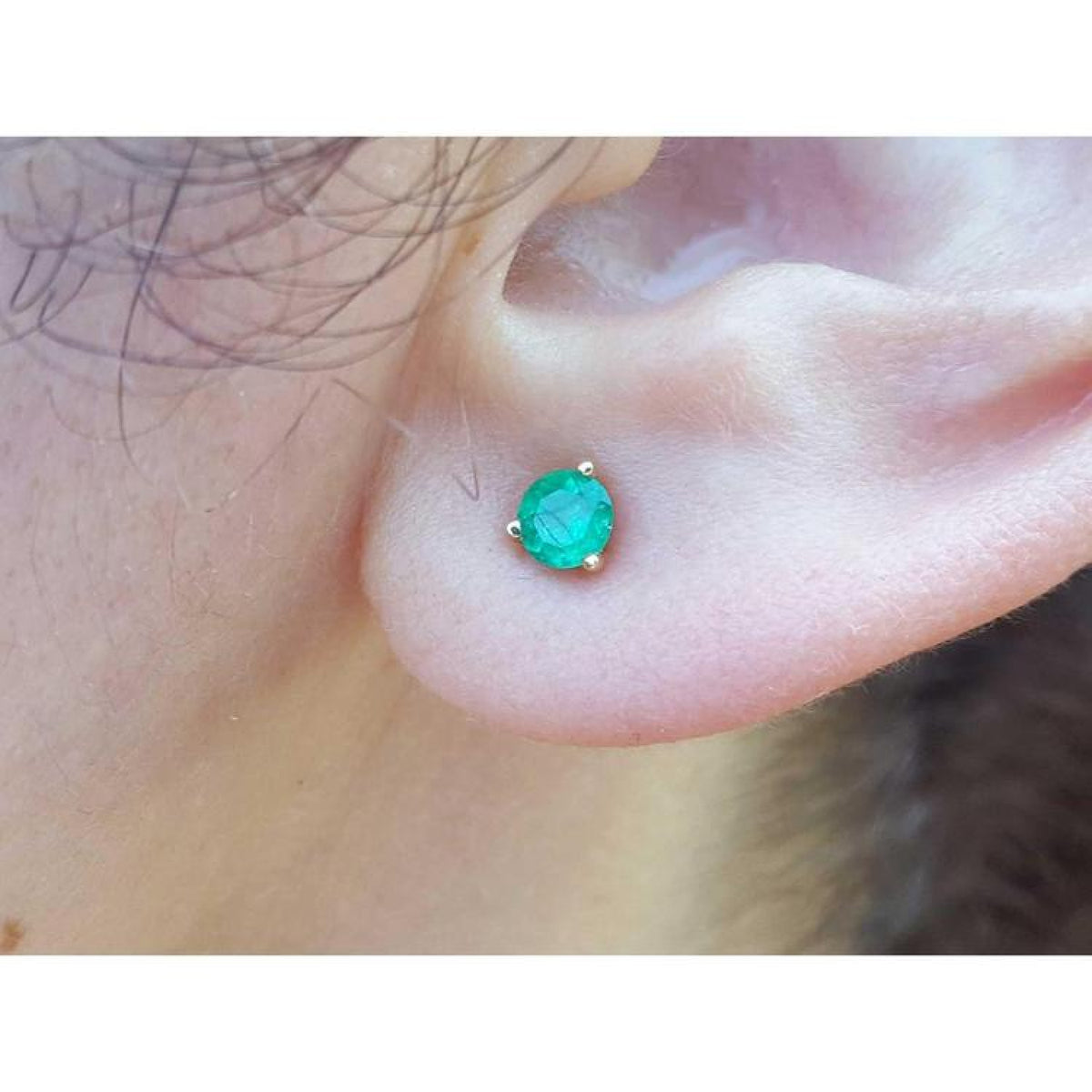 Medium deep green emerald stud earring in 14K yellow gold with three prongs, worn on an ear.