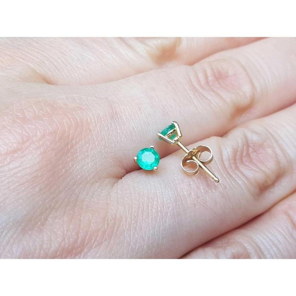 Round medium deep emerald stud earrings in 14K yellow gold with three prongs, held on a hand.