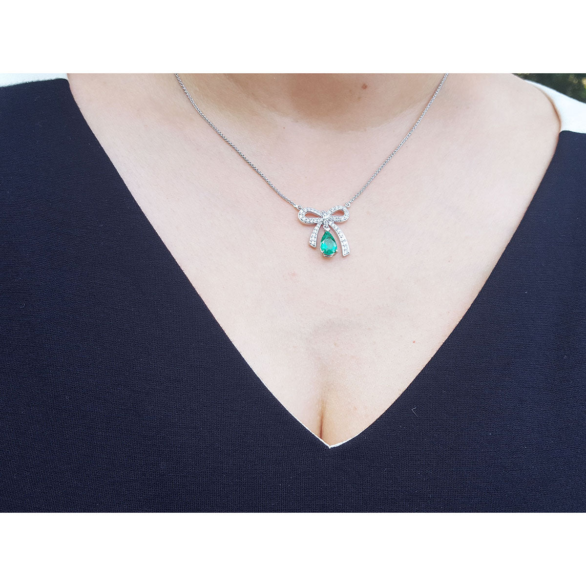 18k white gold bowtie necklace with a Colombian emerald pendant worn by a lady in a navy blue top.