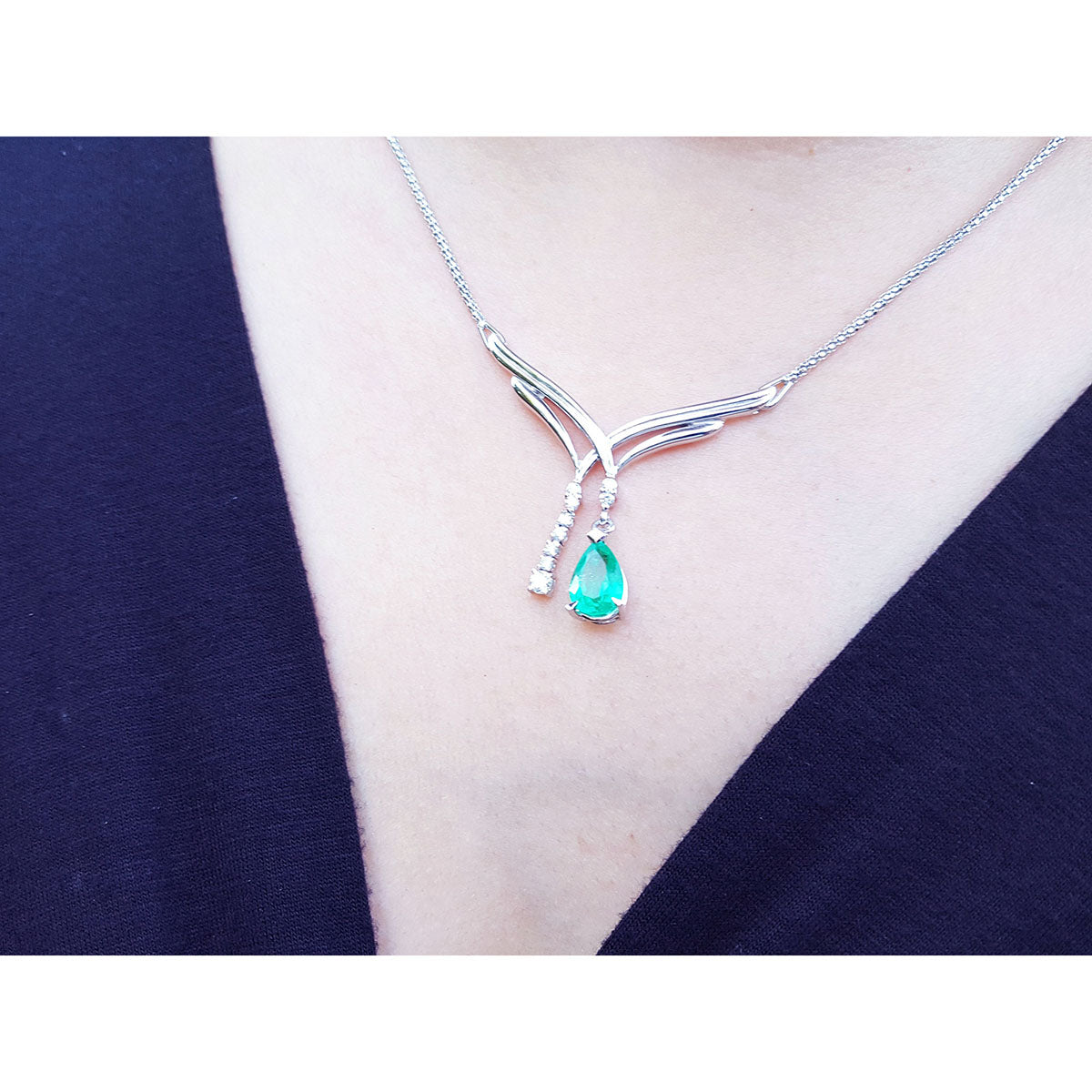 A teardrop emerald necklace with a green teardrop pendant on a person wearing a dark blue top.