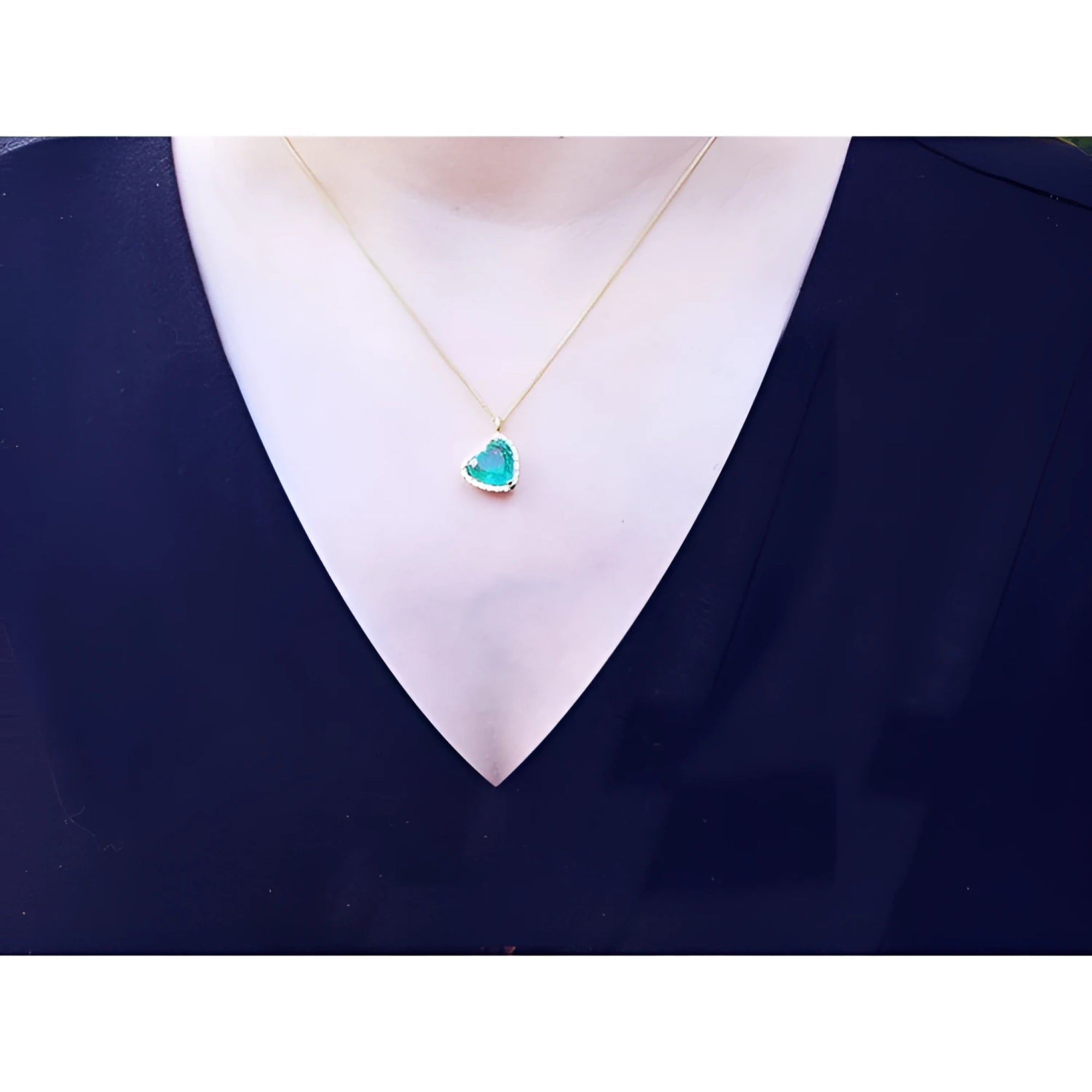 Tilted heart emerald necklace with halo diamonds on an 18K gold chain, worn on a woman's neck.