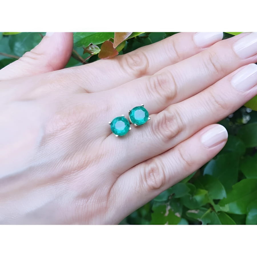 Round bluish-green Colombian emerald stud earrings in 14K yellow gold on a hand.