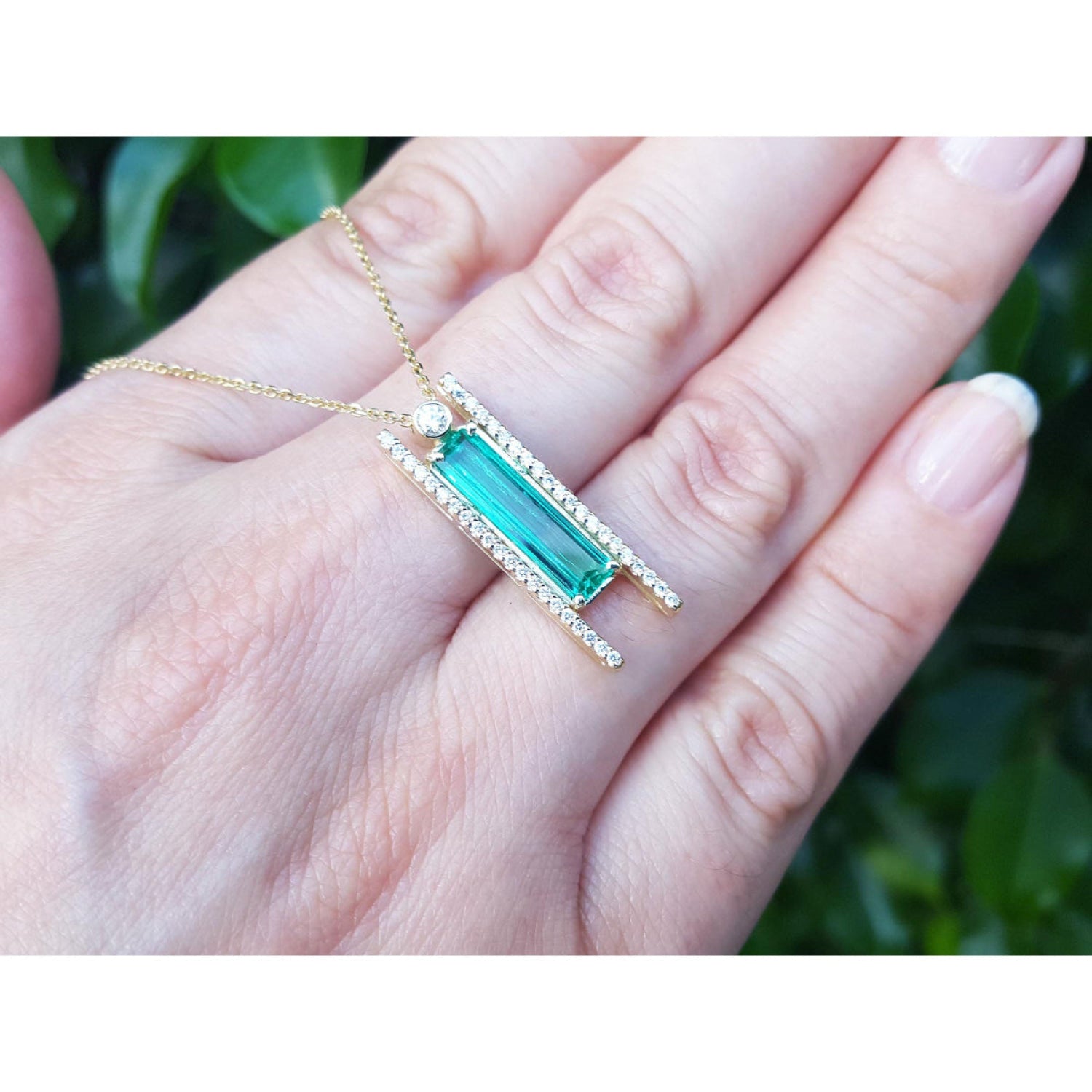Emerald baguette cut necklace with natural diamonds, 14K yellow gold, and sliding tube bail chain. shown resting on a person's hand against a blurred green background.