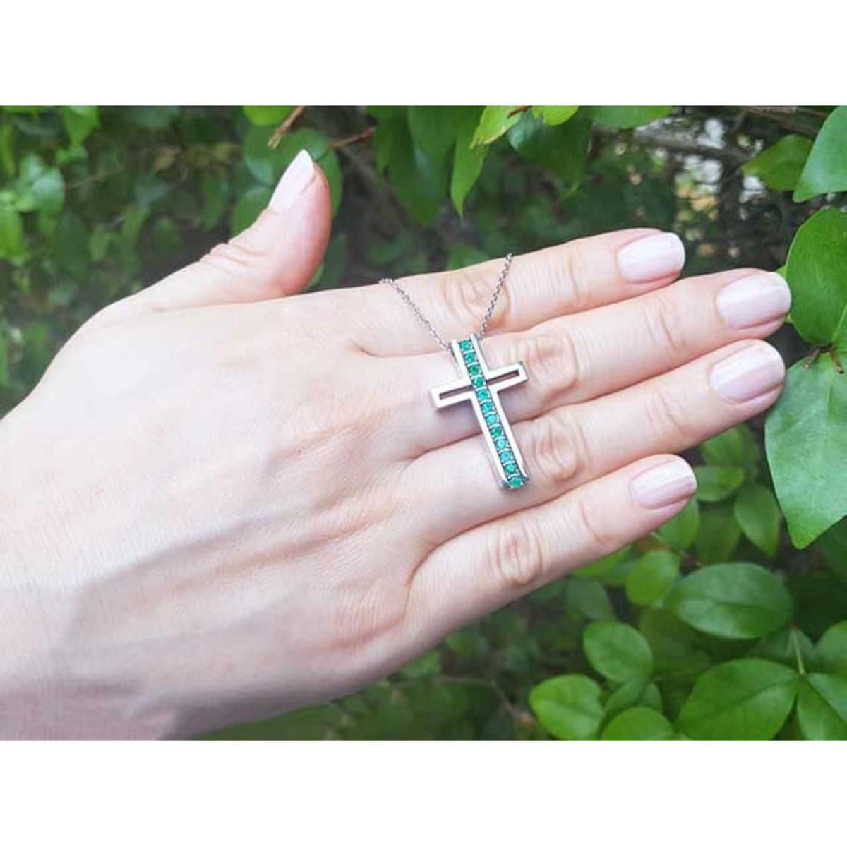 Hand holding a white gold cross necklace with natural emeralds against a green leafy background.
