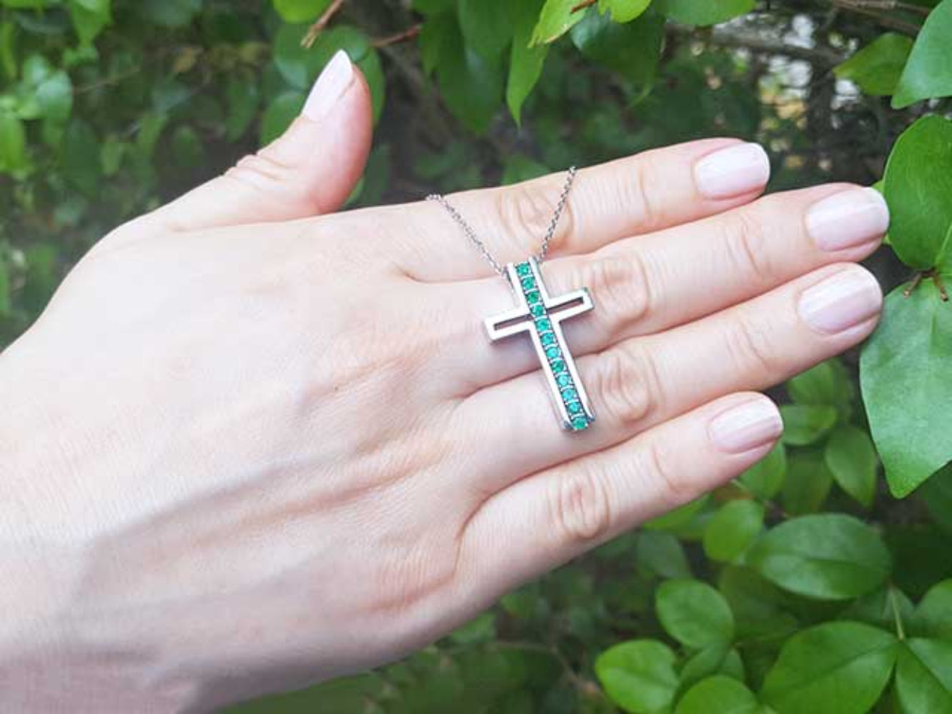 Hand holding a white gold cross necklace with natural emeralds against a green leafy background.