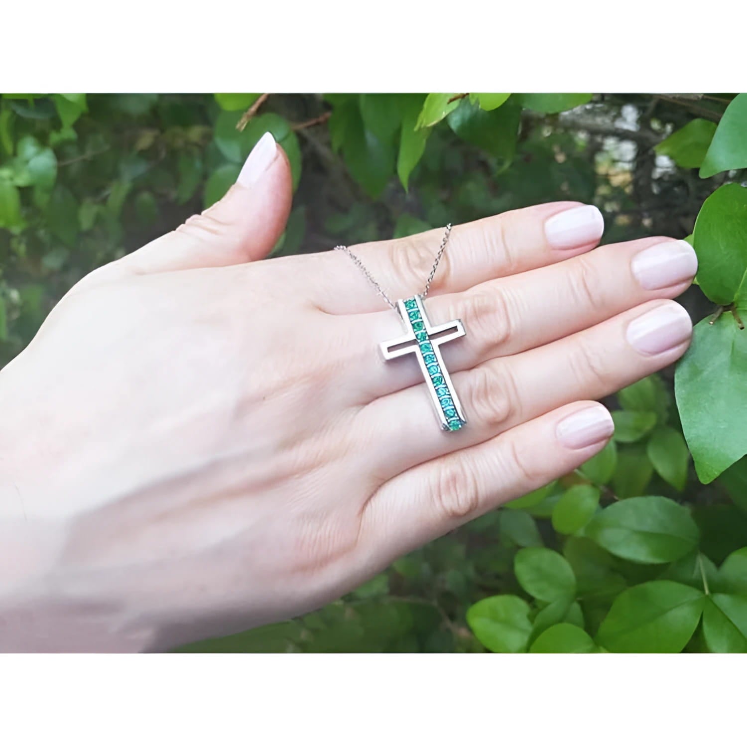 14K white gold cross pendant with prong-set round Colombian emeralds on a delicate chain, held in a hand.
