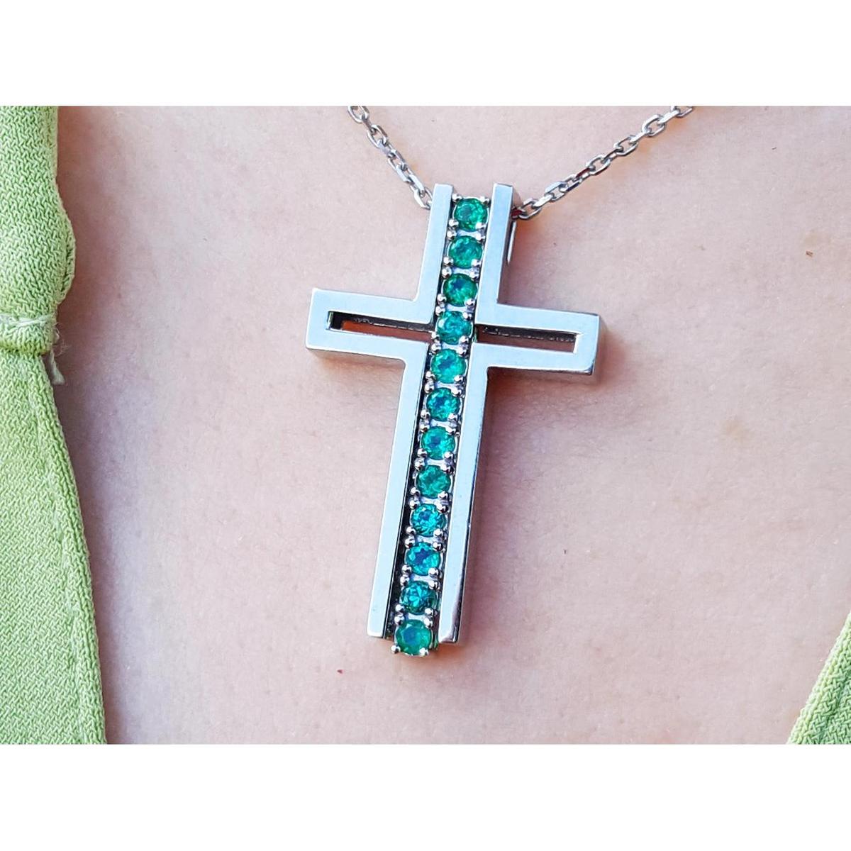 A white gold cross pendant with round cut natural emeralds on a chain, worn on a person's neck.