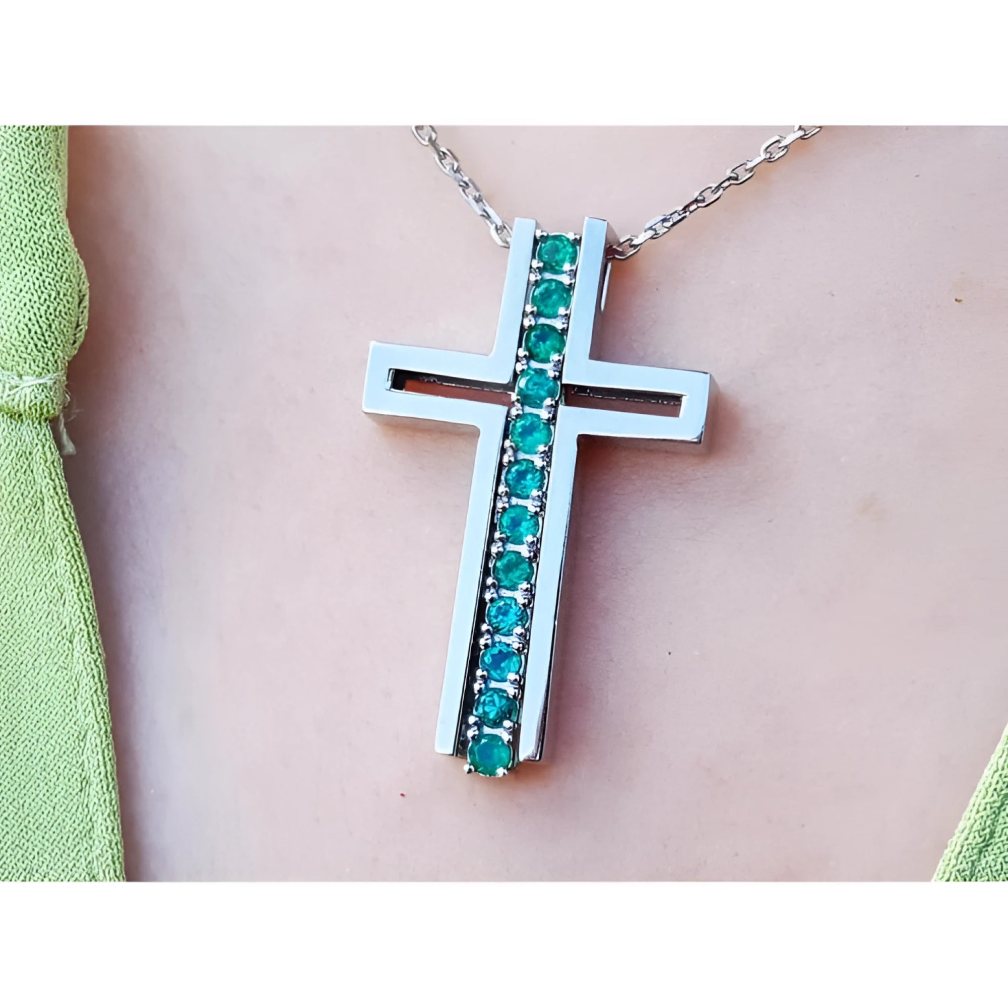 A white gold cross pendant with round cut natural emeralds on a chain, worn on a person's neck.